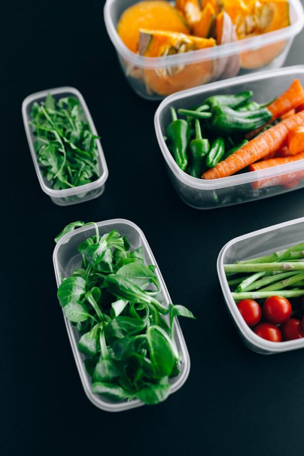 Assorted fresh vegetables including arugula, carrots, and tomatoes in plastic containers against a black background.